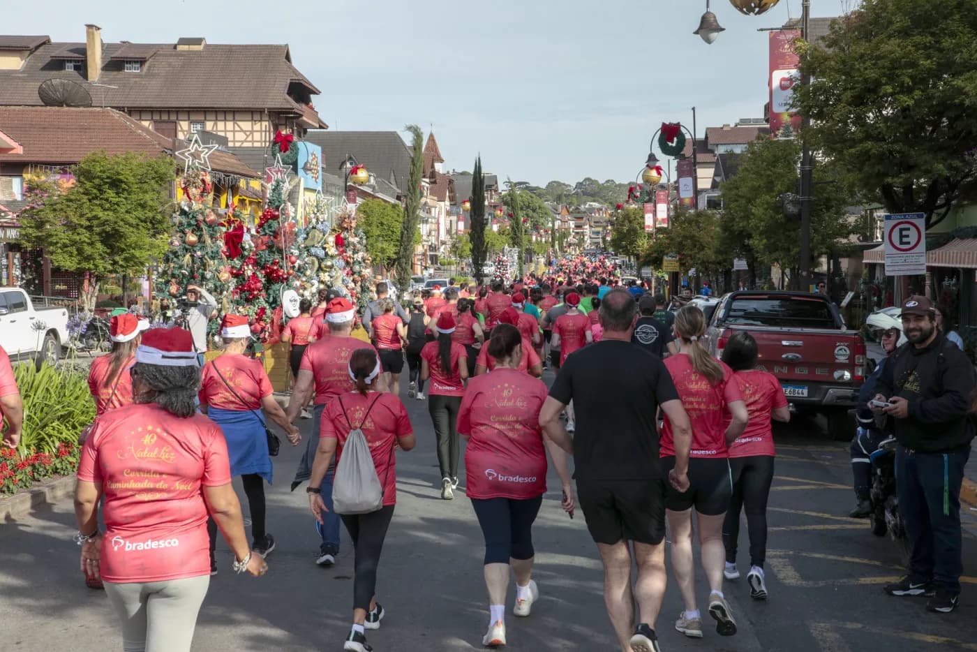 Participantes de gorro vermelho caminham pela avenida ao lado de árvores de Natal decoradas.