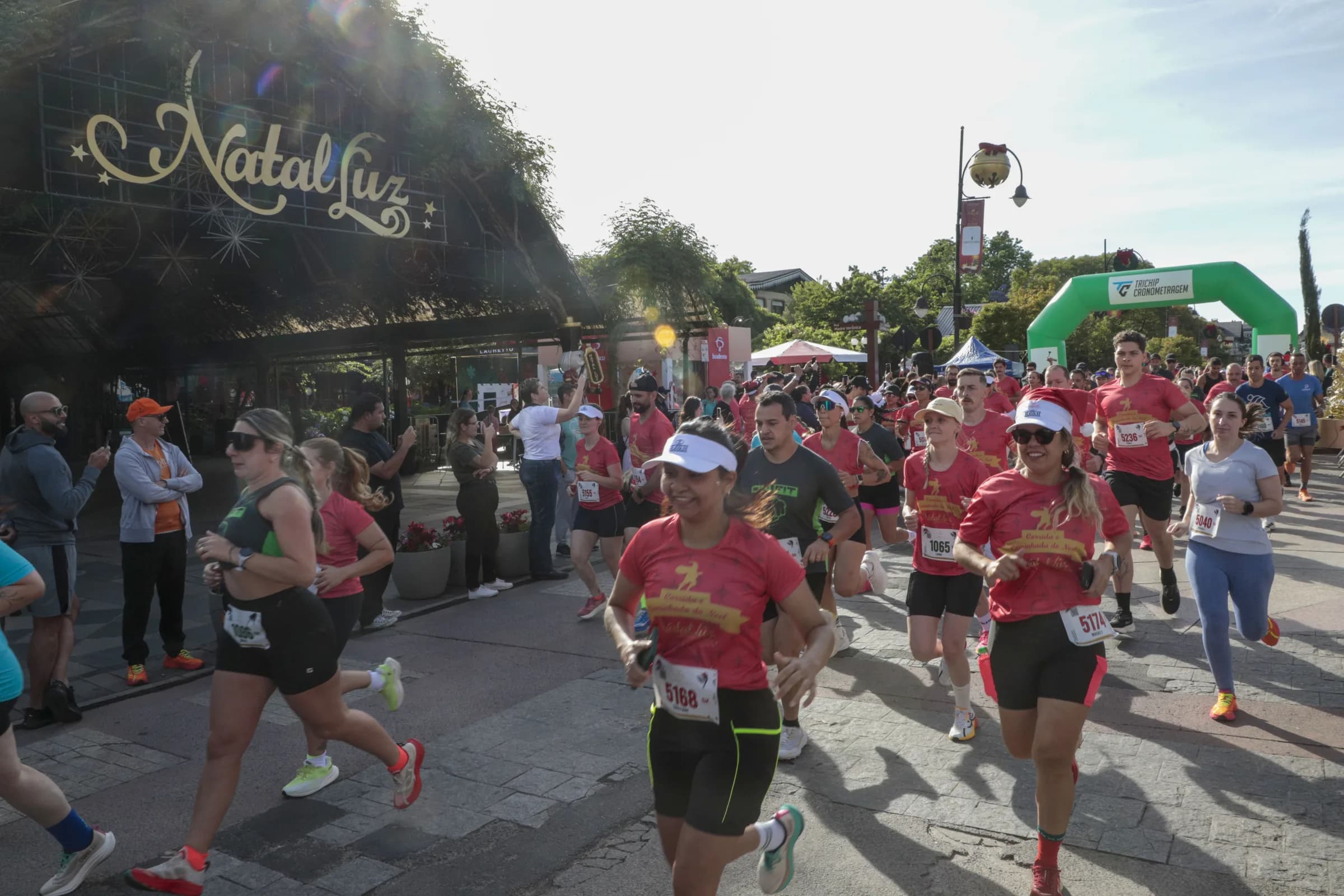 Largada da Corrida do Noel na Rua Coberta: corredores de camiseta vermelha passam sob o letreiro do Natal Luz, com o pórtico de cronometragem ao fundo.