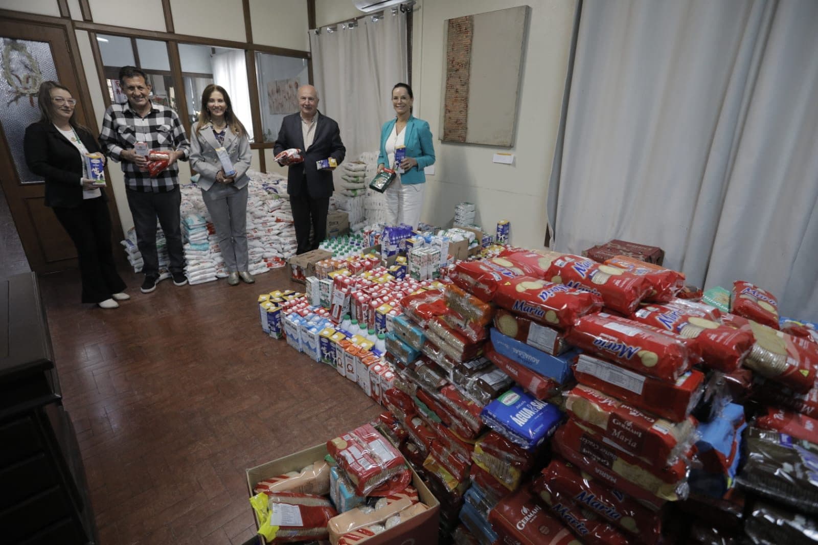 Cinco pessoas seguram produtos em uma sala repleta de alimentos arrecadados, como pacotes de biscoito, caixas de leite e sacos de arroz e açúcar.