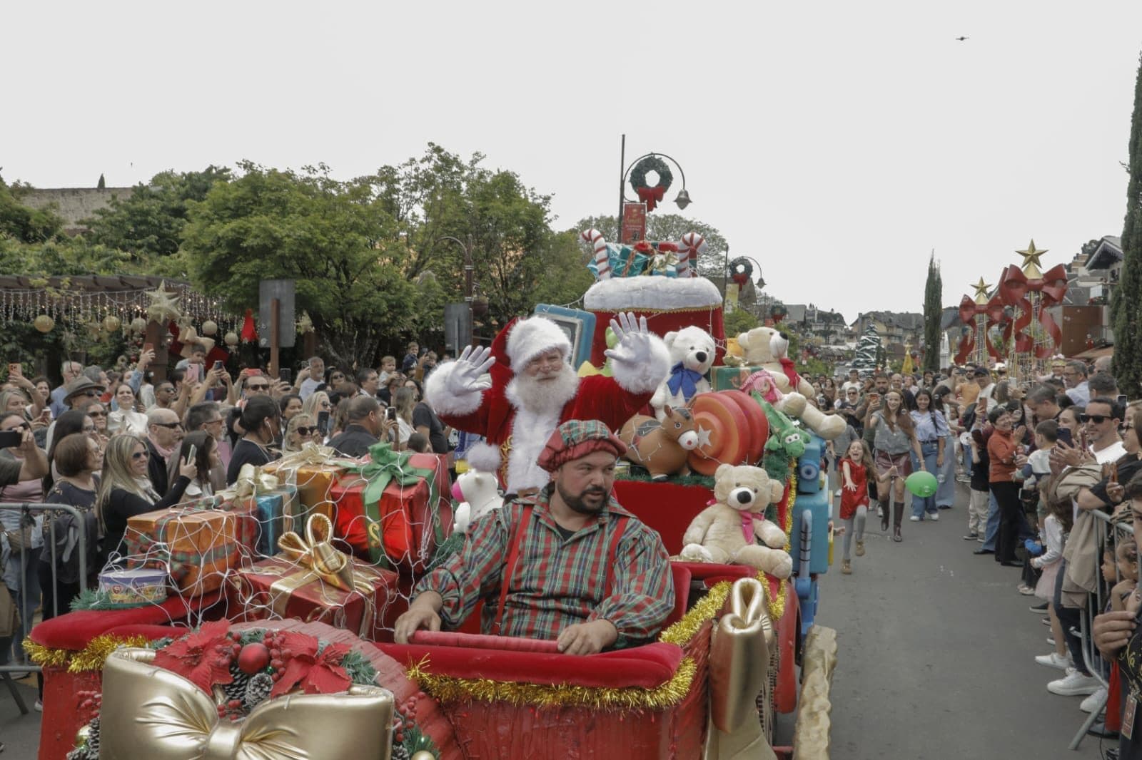 Papai Noel acena do alto de um carro alegórico decorado com presentes e ursos de pelúcia, enquanto o público lota as calçadas da avenida durante a Parada de Natal.