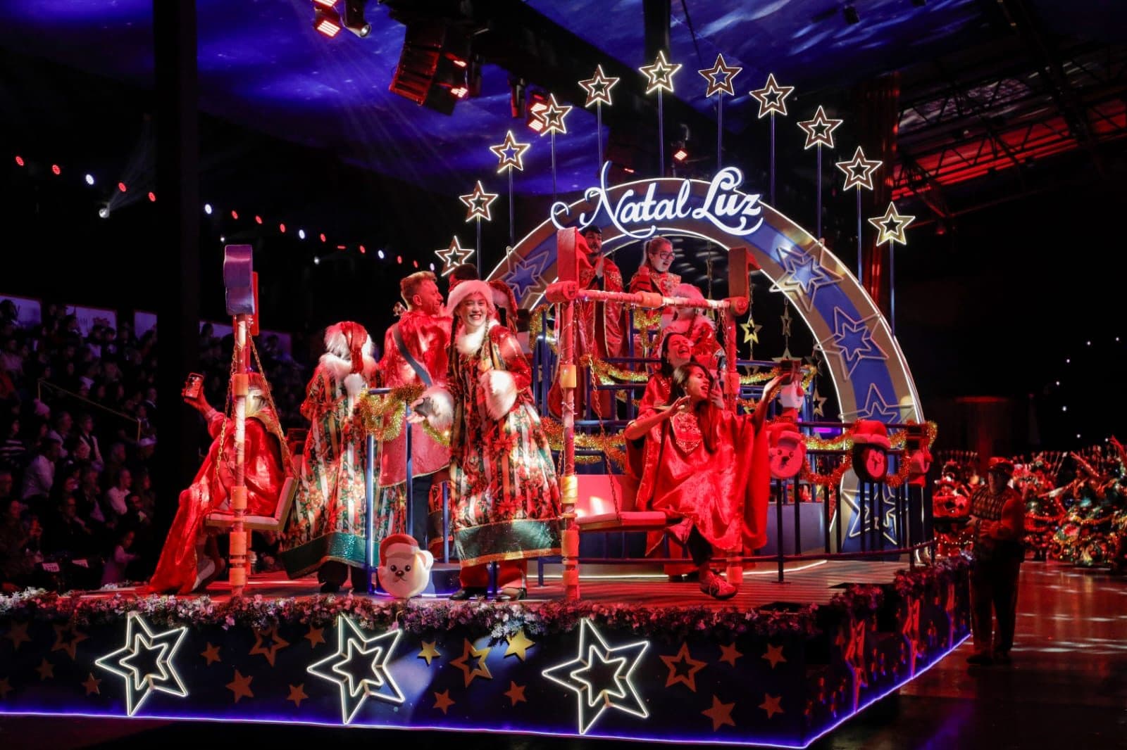 Participantes com figurinos natalinos vermelhos desfilam sobre carro alegórico iluminado com estrelas e arco com o letreiro Natal Luz, diante da plateia.