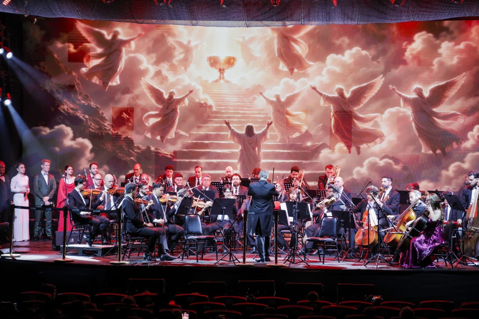 Orquestra sinfônica se apresenta no palco sob a regência de um maestro, com cantores ao lado e um telão ao fundo exibindo anjos entre nuvens e uma escadaria iluminada.