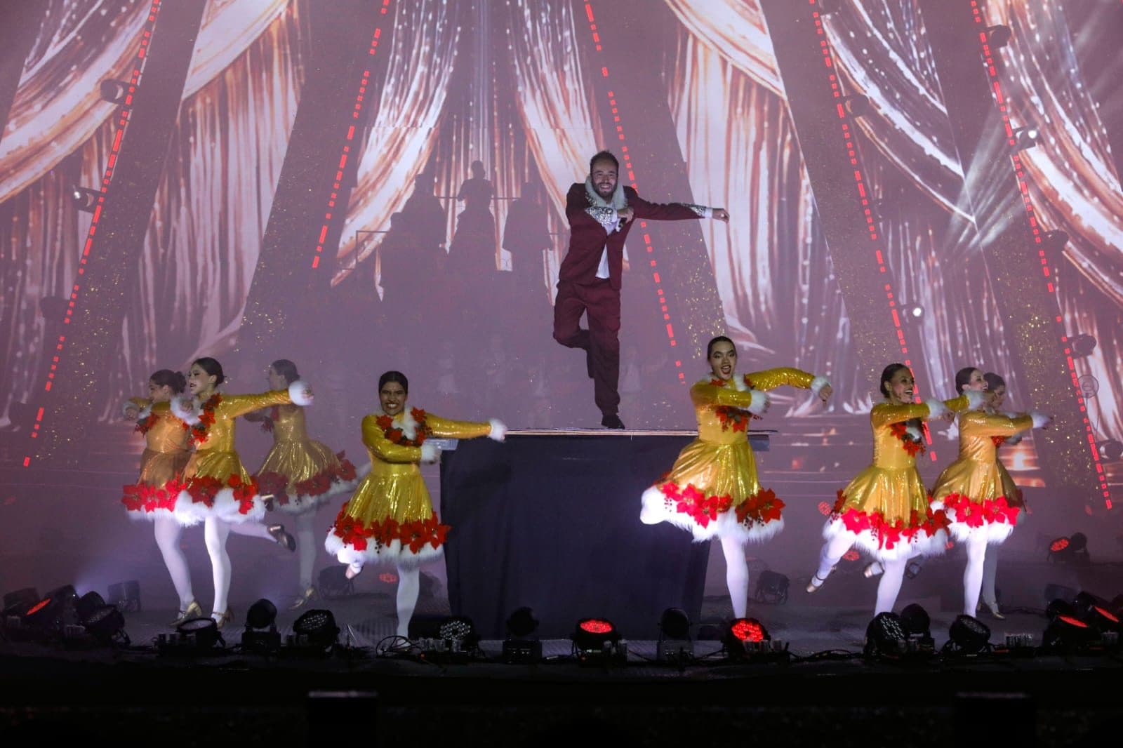 Solista de terno vinho dança sobre um praticável no centro do palco, cercado por bailarinas com vestidos dourados e barras de flores vermelhas, diante de um telão com cortinas iluminadas.